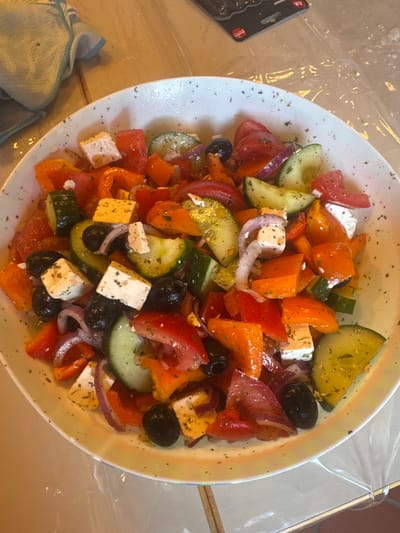 A white ceramic bowl filled with a Greek salad sits on a reflective, light-colored surface, possibly a table covered with plastic wrap. The salad contains chopped tomatoes, cucumbers, red onion, orange bell pepper, black olives, and cubes of feta cheese. The ingredients are mixed together and appear to be seasoned with herbs. In the upper left corner of the image, a folded light blue towel with a white and blue trim is visible. The background is out of focus, with hints of wood grain. The image is taken from a slightly elevated angle, looking down into the bowl of salad. There are no people or visible actions depicted. The location in Merzenich, Germany, is not visually represented in the image.
