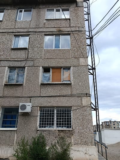 The image shows a close-up, low-angle view of the exterior of a multi-story building with a rough, pebble-dash concrete facade. The building appears to be in a state of disrepair. On the right side of the building, a black metal fire escape or external ladder runs vertically. A bundle of thick electrical wires is strung along the side of the building and across the sky.
The windows on the building are of various types and conditions. Some have clear panes, while others appear to have broken or covered glass. One window has an orange, opaque material covering a portion of it. An air conditioning unit is mounted on the wall beneath one of the windows. Below the air conditioning unit and to its right are two windows, one with a metal grid, and the other showing blue storage bins with labels.
In the foreground, at the bottom of the image, is a dense green bush. In the background, across a paved area with a white wall and a railing, other apartment buildings are visible under a cloudy sky. The overall impression is of an older, functional but weathered urban environment. This building is located in the city of Satbayev, Kazakhstan, a city known for its mining industry.