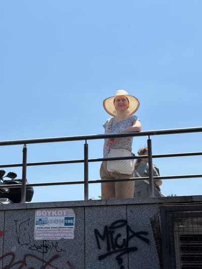 A woman wearing a wide-brimmed straw hat, glasses, a patterned blouse, and khaki shorts leans on a railing against a clear blue sky. She is carrying a white crossbody bag with a pattern. Behind her, another person in a green top is partially visible. The railing is a dark metal structure with horizontal bars. Below the railing, a concrete wall is covered in graffiti, including the word "BOYKOT" and other illegible text in Turkish. A motorcycle is parked to the left of the frame. The scene appears to be in an outdoor urban setting. The presence of graffiti and a boycott poster suggests a public space possibly in Fatih, Turkey, given the Turkish language on the poster.