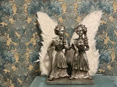This is a medium shot, eye-level photo of two silver angel figurines with white feathered wings, positioned in front of a damask-patterned wallpaper. The wallpaper has a distressed, antique look with swirling motifs in shades of blue, grey, and beige. The figurines are detailed, with intricately designed clothing featuring floral patterns and trim. Their faces are serene, with closed eyes, and they appear to be in a gentle pose, possibly interacting with each other. The overall scene evokes a sense of elegance and a touch of the ethereal. The figurines are placed on a smooth, light blue surface that reflects the light. The lighting appears to be from an indoor source, casting soft shadows. There is no visible text in the image. The context of Bielefeld, Germany, does not provide specific visual cues within the image itself.
