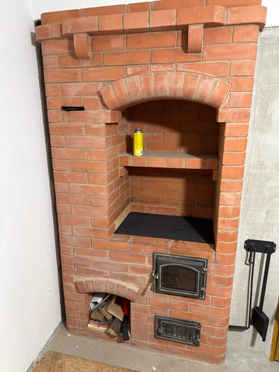 This is a full-shot, eye-level, indoor photograph of a red brick oven in a room. The oven is tall and narrow, built from red bricks with light grey mortar. It has an arched opening at the top with a shelf inside. A yellow can of spray paint sits on the shelf. Below this is a black stovetop with two circular burners. Further down, there are two metal doors on the oven. The upper door has a glass window and decorative trim. The lower door is smaller and has a slotted vent. To the left of the oven, a small arched opening contains firewood and an axe with a red handle. To the right of the oven, a black fireplace tool set with a shovel and poker hangs on the wall. The floor is a light-colored concrete with a patterned rug in the lower left corner. The walls in the background are a plain white and concrete. The oven appears to be in a state of construction or recently built. There are no people in the image. The location is stated to be in Пятрышкаўскі сельскі Савет, Belarus.