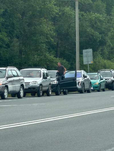 This image, taken in Novaya Bykovka, Russia, shows a line of vehicles parked along a road. The vehicles appear to be SUVs or similar passenger cars. One person, a man wearing a dark t-shirt, is standing by the open driver's side door of a dark-colored SUV. He is leaning into the car and appears to be interacting with someone inside. Other cars in the line are also visible, including a light-colored SUV with a child visible in the backseat, and a green vehicle further down the line. The scene is set against a backdrop of lush green trees, suggesting it is daytime and the weather is likely pleasant, possibly overcast. The road has white lines marking the lanes. A signpost with a reflective surface and a small yellow element is visible in the background. The overall impression is of a brief stop or a queue of vehicles.