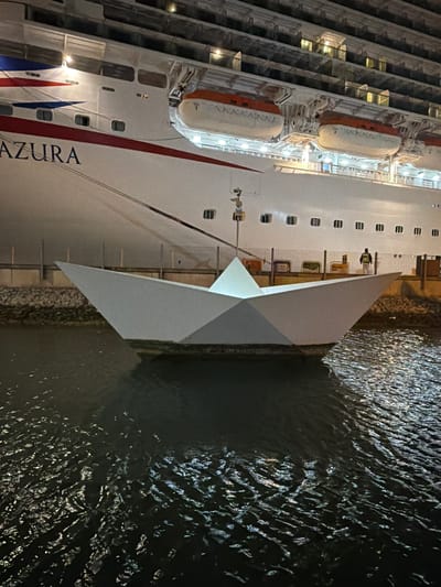 The image displays a large cruise ship, identified by the partially visible name "AZURA" on its hull, docked at a port during nighttime in Floriana, Malta. The ship is brightly illuminated, with lights visible from numerous windows and decks, contrasting with the dark surroundings.

The white hull of the ship features multiple stacked decks, many with their interior lights on, suggesting occupied cabins or balconies. Several orange and white lifeboats are suspended along the upper sections of the hull. Towards the bow, distinctive red, white, and blue stripes are painted on the ship's side. Below the ship, a calm body of water reflects the vessel's lights, showing subtle ripples across its dark surface. A concrete or stone pier separates the ship from the water.

A lone individual, possibly a dock worker or crew member, is visible on a lower deck or the dock, wearing a high-visibility vest, indicating a functional rather than leisure-oriented moment. The overall scene conveys the quiet activity of a large vessel moored in a harbor after dark.