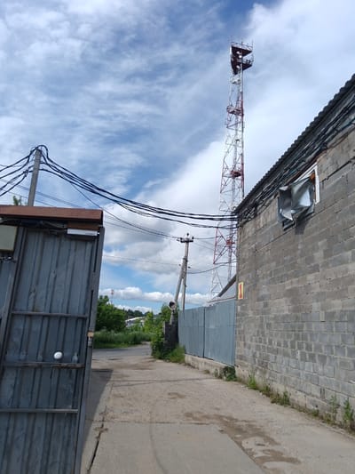 The image depicts a street scene in the city of Bogoslovo, Russia. On the right, a gray cinder block building with a corrugated metal roof is visible. The building has a section of its wall where the metal sheeting is bent outwards from a window opening. A tall, lattice radio tower, painted white and red, stands in the background, piercing a cloudy blue sky. To the left, a large, gray corrugated metal gate is partially open, revealing a glimpse of an interior space. Several electrical poles with numerous black wires crisscross the sky above the street and buildings. A gray metal fence runs along the side of the cinder block building. The ground in the foreground is paved with asphalt and concrete. Green trees and vegetation are present on either side of the street, suggesting an outdoor, possibly industrial or semi-industrial, area.
