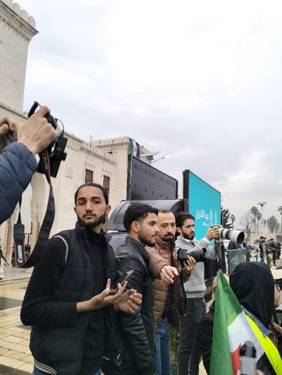 A group of men stand outdoors on a cloudy day, several holding cameras. One man in a black vest holds a smartphone. Another wears a black leather jacket and a third a brown puffer jacket. One man aims a camera with a large lens. A flag with green and white stripes is visible in the foreground. A building is in the background, and a Syrian flag is partially visible on top of the building.