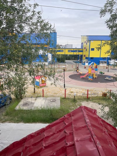 This is a ground-level, slightly elevated, vertical photograph taken in Pangody, Russia. The image captures an outdoor playground area adjacent to a brightly colored yellow and blue building. The building, which appears to be multi-story, has blue window frames and a blue roof. The playground features a variety of colorful equipment, including a red slide, swings, and other climbing structures, all situated on a dark-surfaced play area marked with white lines. A small green toy car is visible on the playground, as is a bicycle with a rider on it. Yellow fencing surrounds parts of the playground and extends around a sandy area.
In the foreground, on the right side of the image, a portion of a red, corrugated metal roof is prominent, obscuring some of the background. Green grass grows around concrete slabs with circular manhole covers. The sky appears overcast, suggesting a cloudy day. Trees with green leaves frame the scene, particularly on the left and top portions of the image. The overall impression is a residential or communal outdoor space, likely in a town or city. The time of day is difficult to ascertain definitively due to the overcast conditions, but it appears to be daylight. No discernible text is visible in the image. The scene conveys a sense of everyday outdoor activity and community space.
