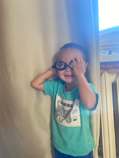 A young child with dark hair and wearing a teal t-shirt with a "Gaming" graphic and blue shorts is looking at the camera while holding goggles over their eyes with their right hand. They are standing next to a light-colored curtain, with a white radiator and what appears to be a stack of boxes or books visible in the background. The image is brightly lit, with some glare. The location is not specified in the image.
