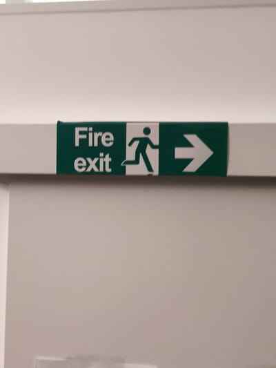A fire exit sign is displayed horizontally on a white wall, above a white door. The sign features a green background with white text and symbols. On the left side of the sign, the words "Fire exit" are printed in white, with "Fire" above "exit." To the right of the text is a white silhouette of a person running, with a circle for a head. Further to the right, a white arrow points to the right. The sign is affixed to the wall with green tape on the left and right sides, which are partially visible. The overall scene appears to be indoors, likely in a building or hallway, given the presence of a door and a fire exit sign. The lighting suggests it is daytime, with no visible weather cues. The location is identified as Troisdorf, Germany.