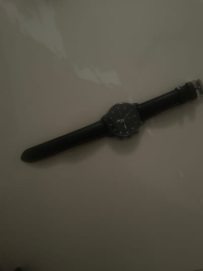 The image shows a black wristwatch with a leather strap lying on a flat, light-colored surface. The watch face displays the time, with numerals at each hour. The hour and minute hands are visible, along with a second hand. The image is taken from a high angle, showing the entire watch and a portion of the surface on which it rests. The location is not Ebo, Nigeria; the scene is indoors on a surface with no geographical markers.