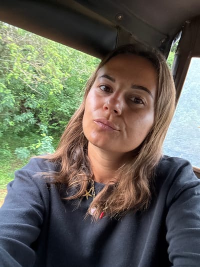 A woman angles her face towards the camera with a slight pout, exhibiting fair skin and brown hair. She wears a black shirt and a thick gold chain necklace with charms including a starfish, a red lip, and a pearl. The background reveals foliage through a window. Part of the vehicle’s interior, including a black panel, is visible.