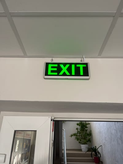 The image shows a hallway with a white ceiling and white walls. A green illuminated "EXIT" sign is mounted on the wall. Below the sign, a doorway leads to a staircase with plants. The scene is likely indoors, possibly in a public building in Nisporeni, Moldova, given the context. No people are visible in the image.