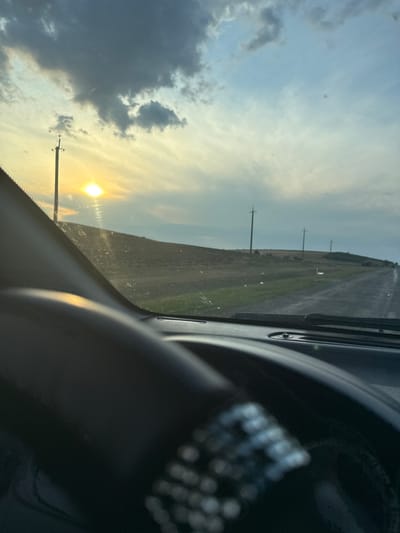 The image is taken from the perspective of a driver inside a vehicle, looking out through the windshield. The scene depicts a rural landscape at dusk or dawn, with the sun low on the horizon casting a warm glow. The sky is partly cloudy, with dramatic formations of gray and white clouds contrasting with patches of clear blue. In the foreground, a portion of the car's dashboard and steering wheel is visible, with some decorative elements on the steering wheel.
The landscape outside consists of rolling hills, with fields of dry grass or crops. A dirt or gravel road stretches into the distance, bordered by more open land. Several utility poles with wires are spaced along the roadside, indicating the presence of infrastructure in this remote area. There are no people visible in the image, and the overall impression is one of solitude and a journey through a quiet, open environment. The location is specified as Iordanovca, Moldova.