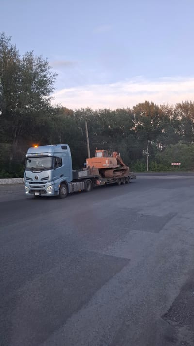 A light blue semi-truck is transporting an orange excavator on a flatbed trailer. The truck is on an asphalt road, and trees line the left side of the road. The sky is clear with some wispy clouds, suggesting it is either early morning or late evening. There is a sign with red and white circles on a pole in the distance. The truck has its headlights on, and the excavator appears to be old and well-used. The scene is likely in Achinsk, Russia, given the location context provided. There is no discernible text on the excavator or trailer, but the truck's license plate is partially visible and appears to be a Russian license plate. The overall mood of the image is neutral, with the focus being on the transportation of the heavy machinery.