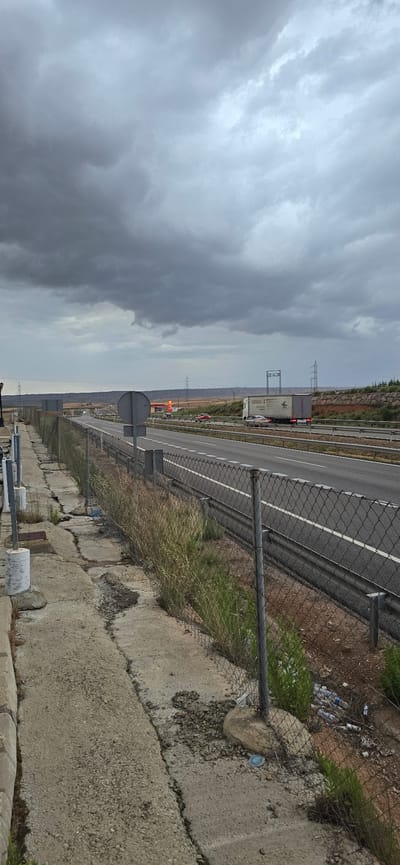 The image depicts an overcast, cloudy day on a highway, likely near Venta de la Romera, Spain. A multi-lane highway stretches into the distance, with traffic including several cars and a large semi-trailer truck. A chain-link fence runs along the side of the highway, separating it from a cracked concrete path and overgrown vegetation. There are also power lines and wind turbines visible in the background, suggesting a rural or semi-rural location. Litter, including plastic bottles, is present along the fence line. The overall impression is one of a functional but somewhat neglected roadside environment under a somber sky.