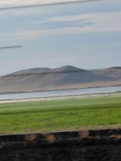 The image depicts a landscape scene. In the foreground, there is dark soil. Behind the soil is a bright green field. Beyond the field is a body of water, likely a lake or river, which appears calm and reflective. Further in the background, there is a large, undulating hill or mountain range. The sky above is pale blue with wispy clouds. The overall scene suggests an open, rural or natural environment. The image does not provide any specific landmarks or details that would allow for identification of the city of Молодёжный, Kazakhstan.