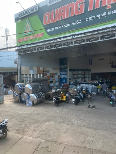 This image depicts the exterior and partial interior of a commercial establishment located at DC 05 - 09 Trần Phú, Xuân Long, Cao Lãnh District, Đồng Tháp Province, Vietnam. The building features a prominent sign identifying it as "QUANG THỊ," advertising "GẠCH MEN • SƠN NƯỚC • THIẾT BỊ VỆ SINH" (tiles, water paint, sanitary equipment), with additional branding for "CEMCOATS" and "ANH VIỆT UC PAINT."
The foreground consists of a concrete ground area. On the left, a stack of multiple large, cylindrical stainless steel water tanks, some labeled "QUỐC THẠNH," are horizontally arranged. A single square tile lies on the ground nearby. A corrugated metal sheet leans against the building's wall.
Under the building's open-fronted entrance, several motorbikes are parked: a yellow scooter, a dark-colored scooter with a wooden pallet strapped to its rear, and a blue electric scooter on the far right. A two-wheeled hand truck is observed lying on its side in the central area.
The visible interior of the store displays walls covered with various square tiles of different colors and textures. A vertical blue banner with "DEXPO" and "QUANG LƯU" text is positioned on the left inside. Shelving units on the right are stocked with numerous bottles, likely paint products. Several potted plants with yellow flowers are arranged near the interior entrance on the right. Two male individuals are present inside; one, wearing a dark shirt and light pants, stands near the potted plants with his back to the viewer, and another is partially visible further in the background. Overhead power lines and a distant telecommunications tower are discernible against the bright sky in the upper left background.