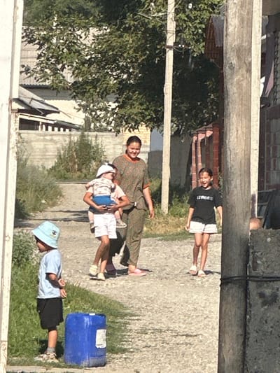 A medium shot captures a sunny, outdoor scene on a dirt path in a residential area of International, Kyrgyzstan. In the foreground, a young boy wearing a light blue t-shirt, black shorts, and a light blue bucket hat stands facing left. To his right and slightly behind him is a large blue plastic barrel.
Moving further back and to the center, a woman in a patterned green and brown tunic is walking, carrying a child on her hip. The child is wearing a white hat, a white and patterned shirt, and white shorts. Behind them, a younger girl in a black t-shirt with white lettering and white shorts walks towards the viewer, smiling and with her mouth open.
The background consists of buildings with some foliage and trees. A light-colored wall with a fence is visible behind the children in the center. A weathered wooden utility pole stands on the right side of the frame, with wires running between it and another pole to its left. The ground is a mix of dirt, gravel, and patchy green grass. The lighting suggests it is daytime, with bright sunlight casting shadows.