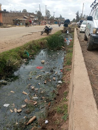 This image depicts a roadside scene in the Tala ward, Kenya, on an overcast day. In the foreground, an open drainage ditch, approximately 1.5 meters wide, runs parallel to a concrete curb on its right. The ditch is heavily polluted with stagnant, dark green water and a dense accumulation of refuse, including numerous plastic bottles (clear, brown), plastic bags (red, green, white), pieces of fabric, discarded footwear, and various unidentifiable debris. Clumps of tall green reeds grow from the water and along the left earthen bank of the ditch.

To the right of the concrete curb is a gravelly dirt shoulder where a light-colored van, equipped with a roof rack, is partially visible, parked facing away from the camera. Its front-right tire, bumper, and lower grille are discernible. Further down this shoulder, two additional cars are parked.

To the left of the ditch, beyond its earthen bank, is a paved road that appears cracked and uneven in sections. A dark-colored motorcycle is parked on the dirt shoulder adjacent to the road, angled towards the left. On the road itself, a person riding a motorcycle, carrying a large bundled item on the back, is moving away from the viewer.

Further along the left side of the road, multiple individuals are visible. Two men are walking away from the camera, one in a light shirt and dark trousers, the other in a red shirt and dark trousers. Other people are congregated near small, makeshift market stalls or shops constructed from corrugated metal and timber. Behind these stalls, a row of single-story stone or concrete block buildings with corrugated metal roofs extends into the background. Overhead utility lines and wooden poles are visible against the cloudy sky.