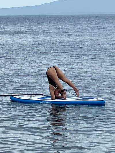 A person is performing a headstand on a blue and white inflatable paddleboard in the ocean. The person is wearing a black swimsuit with white trim. The paddleboard has a leash attached to the person's ankle. The water is calm and there is a distant shoreline with hills. The image is taken from a medium distance, showing the full body of the person and the paddleboard. The location is Dauin, Philippines.
