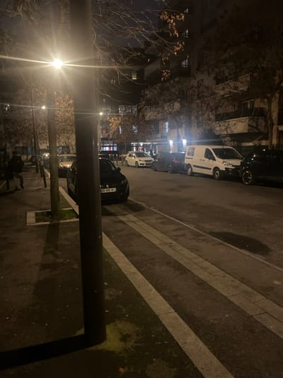 A nighttime street scene with parked cars lining the curb. A lamppost illuminates the foreground. Buildings and trees are visible on the left. The asphalt road features painted white lines. Several people are standing in the left side of the image. A black BMW with visible license plate is parked nearest the lamppost on the right.