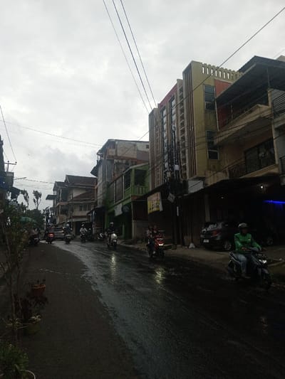 The image depicts an overcast urban street scene in Sindanglaya, Indonesia, characterized by a wet, dark asphalt road curving slightly to the left, indicating recent rainfall. The sky is uniformly grey with visible utility lines traversing overhead. Buildings of varying heights, from two to five stories, line both sides of the street. On the right, a prominent multi-story building features a light yellow-beige facade with vertical architectural fins and a red section near the roofline, adjacent to a structure with balconies and a dark, recessed ground-level area where a dark grey hatchback car is parked. A blue light emanates from within this dark recess. On the far right foreground, a single individual wearing a green jacket and a helmet operates a dark scooter, traveling away from the camera. Further back on the right, two individuals are positioned on a red scooter. Multiple other scooters, some with riders, are visible moving or parked along the street further down the road. Buildings on the left side of the street display a mix of painted and unpainted surfaces, some with visible wear and balconies. Potted plants are arranged along the immediate left edge of the road.