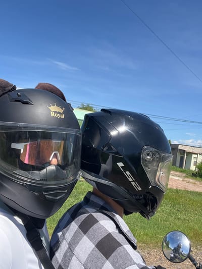 In Hong Son, Vietnam, two individuals, wearing helmets and riding on a motorcycle, are captured in a close-up, forward-facing shot. The person in the foreground wears a black helmet with a gold "Royal" emblem and reflective sunglasses, while the person behind wears a black helmet with "LS2" branding. The background shows a bright blue sky with faint clouds, power lines, and a building with a green roof. The scene suggests a daytime motorcycle ride in a semi-rural or suburban area of Vietnam.