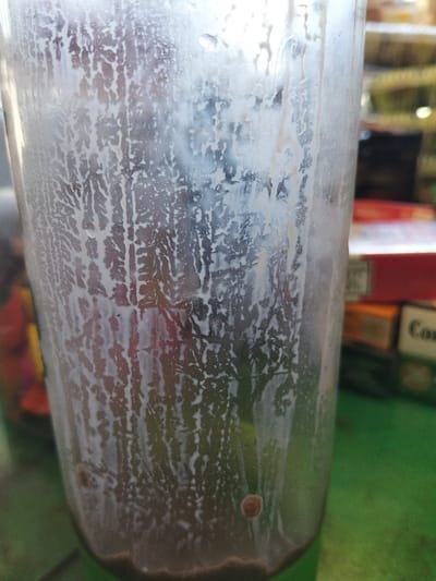 A close-up shows a partially consumed chocolate milkshake in a clear plastic cup. Streaks of milk residue cling to the inside of the glass, creating a dendritic pattern. The remaining milkshake is dark brown. In the blurred background are various objects, including packages of cigarettes. The cup sits on a green surface.