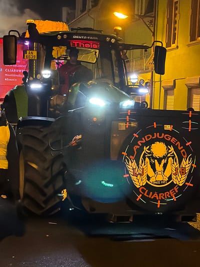 A green Deutz-Fahr tractor with Luxembourgish license plate "AN 6639" is positioned at night on a street in Wasserbillig, Luxembourg. The tractor displays bright LED headlights and additional work lights, along with two amber flashing beacons on the roof. A person in a red garment is visible in the driver's seat. On the tractor's left side, a partially visible sign with a red background lists locations including "CLIÄRRWE," "WAASSERBELLE," and "REIMECH." The name "Theis" is illuminated in red on the front of the cab. A person wearing a yellow high-visibility vest is partially visible on the left side, near the tractor's large rear tire. Attached to the rear of the tractor is a prominent black circular display featuring a yellow and orange logo that reads "LANDJUGEND CLIÄRREF" and "SINCE 1971," with a stylized bull head and wheat sheaves. This display is outlined with reflective orange tape segments. Buildings illuminated by warm yellow streetlights are visible in the background.