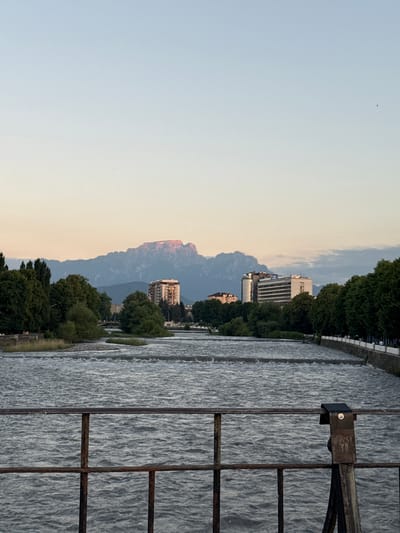 The image depicts a wide, fast-flowing river with a concrete embankment on the right side. A rusty metal railing in the foreground partially obscures the view of the river and the scene beyond. In the middle ground, lush green trees line the riverbanks, with several modern buildings interspersed among them. In the distance, a majestic mountain range dominates the horizon, its peaks illuminated by the warm glow of sunset. The sky is a pale gradient of blue and yellow, suggesting the end of a day. The image appears to be taken from a bridge or elevated vantage point, looking across the river towards the city and mountains. There are no visible people or animals in the scene.
