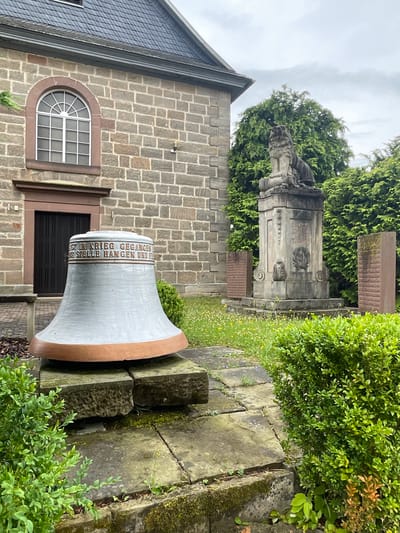 The image depicts a scene outside a stone church in Asterode, Germany. In the foreground, a large, light-colored bell with a copper-colored rim rests on a stone pedestal. Inscriptions are visible on the bell, though the text is not entirely legible. To the right of the bell, a memorial monument stands, topped with a stone lion statue. The monument has inscriptions, including dates that appear to be "1914 - 1918" and the name "DIE GEMEINDE ASTERODE". Flanking the central monument are two smaller, reddish stone tablets with inscriptions. The environment is outdoors, with greenery including hedges and trees, and a cloudy sky overhead. The ground is paved with stone slabs and there are patches of grass and small yellow flowers. The church building, constructed of grey stone blocks with a dark-tiled roof and a prominent arched window, forms the backdrop on the left.