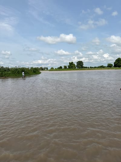 A wide, muddy river flows under a partly cloudy sky in Kunini, Nigeria. On the left bank, a person wearing a white t-shirt and shorts stands in the shallow water near lush green vegetation. The river extends to the horizon, where more trees and vegetation can be seen. The water has ripples on its surface, suggesting a gentle current.