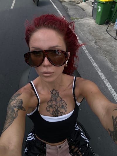 The media file is a low-angle, rotated selfie of a person with bright red hair. They are wearing large, tortoiseshell sunglasses and a black tank top with white trim. The person has tattoos on their chest, left arm, and right arm. They are also wearing black shorts with white stripes and lacing detail, and a light pink jacket is visible. The scene appears to be outdoors, with a paved surface that looks like a road or sidewalk, marked with a white line. In the background, a green recycling bin is visible. The lighting suggests it is daytime. The overall impression is casual and outdoorsy.