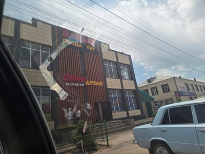 A light blue car is parked on a street in front of a building with signs in Cyrillic. The building appears to be a store, with "БАЛЫ", "СООДА", "УЙ", "СУПЕР", "МАРКЕТИ", and "АРЗАН" visible on its facade. There are people standing outside the store, including a person in a wheelchair. The sky is partly cloudy, suggesting it is daytime. The scene is set in Kyzyl-Suu, Kyrgyzstan.