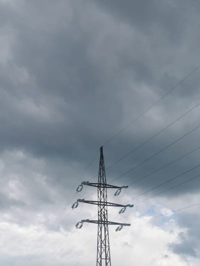 An outdoor, low-angle, full shot features a dark, overcast sky with heavy cloud cover. In the foreground, a tall, dark-colored metal electricity pylon rises from the bottom center of the frame. The pylon is a lattice structure with multiple crossbars supporting power lines. Three sets of insulators, each holding multiple power lines, are visible extending to the right from the pylon. The power lines crisscross diagonally upwards across the frame. The scene is devoid of any people or animals. The environment appears to be an open area with the sky as the dominant backdrop, suggesting a location where such infrastructure is present, possibly on the outskirts of the city of Serov, Russia, given the context. The lighting is dim and diffused due to the cloud cover.