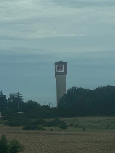 The image captures a tall, rectangular structure with a logo that reads "AJAX" on its upper section, oriented vertically due to the camera's orientation. The structure appears to be a water tower or a similar industrial building. It is situated in an outdoor setting, with a field of dry grass or crops to its right and a dense line of trees to its left. The sky is overcast, suggesting a cloudy day. The overall scene appears to be rural, possibly a former industrial site or an agricultural area. There are no people visible in the image. The time of day is not discernible due to the overcast sky. The location is identified as Bozduganovo, Bulgaria.