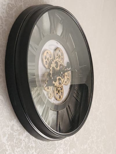 The image shows a close-up, angled view of a black-framed decorative wall clock with Roman numerals. The clock features visible gold-colored gears in its center. The background is a light-colored wallpaper with a subtle damask pattern. The clock is mounted on a wall. The image does not provide any information about the city of Idar-Oberstein, Germany.
