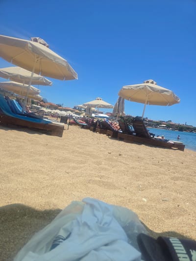 The image captures a sunny day on a beach in the Municipal Unit of Lefktro, Greece. The foreground is dominated by golden sand, with a white plastic bag containing clothing and a pair of black slides partially obscuring the view. In the mid-ground, several beach chairs are arranged under large, beige umbrellas. Some chairs are occupied by individuals sunbathing. The background reveals a clear blue sky stretching over the turquoise waters of the sea. Along the coastline, buildings and more umbrellas suggest a populated tourist area. A few people are visible in the water, swimming or wading. The overall scene depicts a typical summer day at a Greek beach resort.