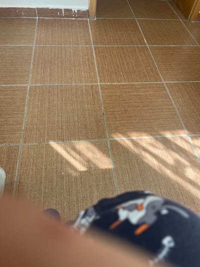 The image shows a close-up view of a tiled floor with diagonal sunlight cast upon it. In the foreground, a blurred portion of a person's leg or arm is visible, wearing dark fabric with a cartoonish pattern of grey, white, and orange figures. A white shoe is partially visible to the left. The tiles are a uniform brown color with thin grey grout lines. The background shows a white baseboard with some white paint drips, and a wooden door frame on the right. The location is identified as Eshmataul, Uzbekistan.