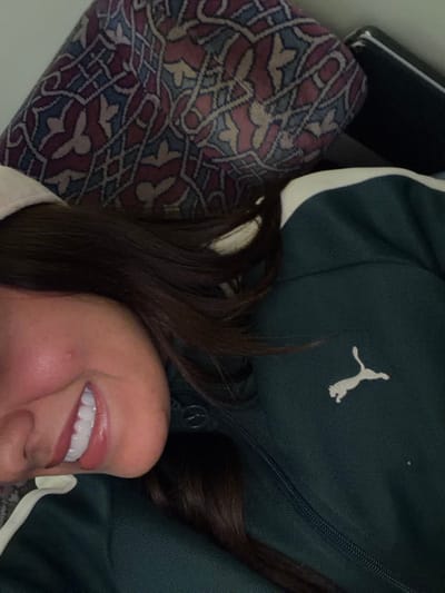 A close-up shot presents a person resting against a patterned cushion, wearing a dark green Puma jacket with a white stripe. Only the side of their face and part of their dark hair are visible. The person is smiling, revealing a set of white teeth. The Puma logo is visible on the upper right side of the jacket. The shot is taken from a slightly elevated angle.