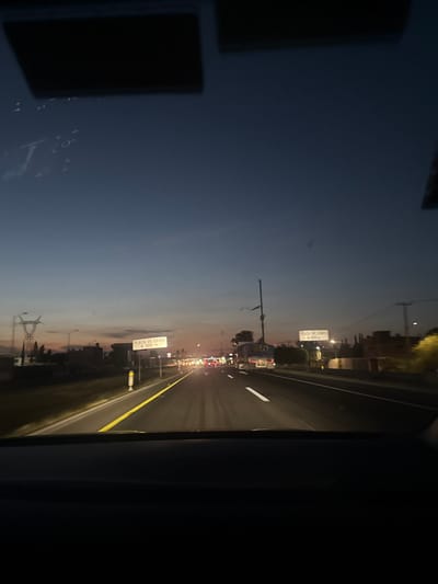 A nighttime highway view from inside a vehicle. The sky is a gradient of dark blue to orange, suggesting dusk or dawn. Multiple light sources are visible, probably from other cars and streetlights. Road signs flank the highway. A yellow line divides the road. The vehicle's interior is visible on the sides, with dark shapes obstructing some view.