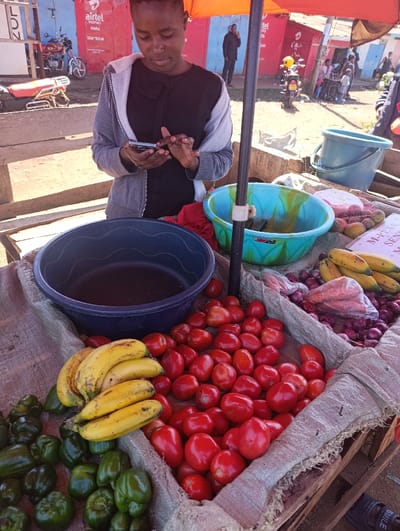 This image captures a vibrant outdoor market scene in Kaplelwo, Kenya, on a bright, sunny day. The central focus is a young woman with short, dark hair, wearing a black top and a grey hooded cardigan, intently engaged with her smartphone. She appears to be operating a market stall, evidenced by the array of fresh produce displayed before her.
The stall, an informal structure likely made of wooden crates lined with burlap, showcases a bountiful selection of vegetables and fruits. Prominently featured are large piles of glossy red tomatoes and vibrant green bell peppers. Alongside these are bunches of ripe yellow bananas, some with slight brown spotting. In the background of the produce, red onions are visible in a clear plastic bag, next to what appears to be carrots in another bag, and some smaller, lighter-colored fruits in a white net bag.
Two plastic basins are part of the stall setup: a large, dark blue one and a smaller turquoise-and-green striped one. A light blue plastic bucket is also visible on a raised surface to the right. An orange umbrella or awning provides shade over the stall.
In the background, other people are visible, including a man standing further back, also appearing to be on his phone. Motorcycles are parked on the dirt ground. The backdrop consists of simple buildings with brightly painted walls, including a prominent red building with an "airtel money" sign, below which "HAPA" and blurred numbers are visible. Another sign partially visible on the right reads "M-PESA SEN...", indicating another popular mobile money service. The overall scene depicts a typical bustling day of commerce in a local Kenyan market.