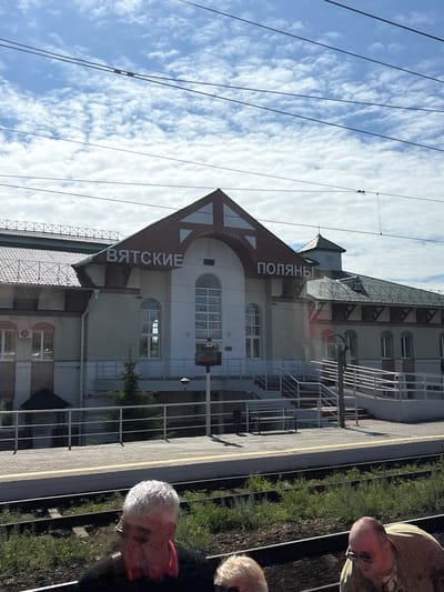 This image captures a train station in Vyatskiye Polyany, Russia, on a bright, cloudy day. The central focus is the train station building, identified by the large white Cyrillic letters "ВЯТСКИЕ ПОЛЯНЫ" (VYATSKIYE POLYANY) affixed to its roofline and upper facade. The building features a prominent central gabled section with a dark tiled roof, light-colored walls (likely beige or light grey), and multiple arched and rectangular windows. A smaller, decorative tower with a pyramidal roof is visible on the right side of the building.

In the foreground, a concrete railway platform runs parallel to multiple railway tracks. The platform is equipped with white metal railings, a set of concrete stairs, and an accessible ramp on the right side. A yellow safety line marks the edge of the platform. A black bench and a black waste receptacle are positioned on the platform. Between the platform and the immediate foreground, several railway tracks with wooden sleepers and gravel ballast are visible, with green grass and weeds growing sparsely between them. A small conifer tree is planted near the platform railing.

Above the station, a light blue sky is populated with numerous white, fluffy cumulus clouds. Several dark electrical or communication wires traverse the upper portion of the frame.

In the bottom foreground, partially obscuring the railway tracks, are the heads and shoulders of three individuals. On the left, a man with short, grey hair and glasses is visible, wearing a dark garment with a red collar or scarf. A portion of his face appears discolored. To his right, only the top of a head with light blonde or white hair is visible. Further right, a man with a bald head and glasses, wearing a light-colored (possibly tan or olive) collared shirt, is looking downwards and to the right.
