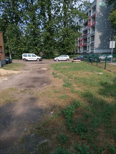 The image depicts an exterior scene in Bytom, Poland, likely in a residential area. In the foreground, a dirt path with patches of dry grass and some green vegetation is visible. To the right, a green metal fence encloses an area with more grass. Beyond the fence, several cars are parked, including a dark sedan and a light-colored hatchback. In the middle ground, a white van is parked on a paved surface, and another light-colored hatchback is visible. A multi-story apartment building with balconies and trees in front of it is prominent in the background. The building has a grey facade with red accents on some of the balconies. The sky is clear and blue, suggesting it is daytime. There are no discernible human figures engaged in any specific activity.