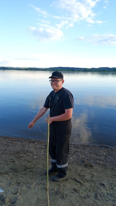 An adult male stands on a sandy and pebbly shoreline adjacent to a large body of water in Шолья, Russia. The man, positioned slightly right of the frame's center, wears a black baseball cap, black-framed glasses, a black t-shirt, and black overalls with grey reflective tape bands on the lower legs, covering black boots. He exhibits a light beard and a slight smile while looking towards the viewer. His right hand grasps a long, slender green stick, acting as a makeshift fishing pole, from which a white fishing line extends downwards. His left hand, extended towards the water, also interacts with the fishing line. The water, calm and dark blue, fills the midground, reflecting the light blue sky with scattered white clouds that dominate the upper frame. Distant forested landmasses are visible on the opposite bank of the water. The ground beneath the man shows footprints in the sand and scattered pebbles near the waterline. The scene is illuminated by natural daylight.