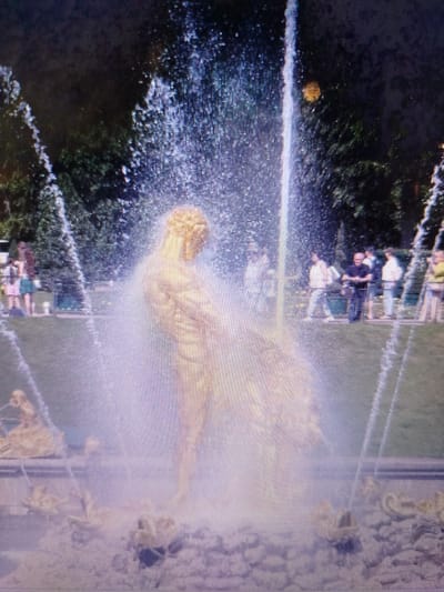 The image shows a golden statue of a muscular male figure, likely a mythological god or hero, as part of a large fountain. The statue is positioned centrally and appears to be the main subject of the fountain's elaborate water display. Water streams upwards from behind the statue and cascades down around it, creating a misty, ethereal effect. The statue itself is illuminated and appears golden, with intricate musculature visible despite the water's obscuring spray.

In the background, a park-like setting is visible with lush green trees and manicured grass. Several people are scattered throughout the background, standing and observing the fountain. They appear to be casually dressed, suggesting a public park or tourist attraction. Some individuals are standing in groups, while others are more isolated. The overall atmosphere suggests a sunny day, with the water from the fountain catching the light.

The statue is surrounded by a stone basin with more ornate sculptural elements at the base, also appearing to be in a golden finish, possibly depicting mythological creatures or decorative motifs. The ground around the fountain is covered with small stones. The image quality is somewhat grainy and appears to be a photograph taken at a distance, possibly with a phone camera.

The location is identified as the city of Snizhne, Ukraine. This suggests the fountain is a landmark or a feature within a public space in that city. The presence of trees, grass, and a fountain indicates a park or a garden area.