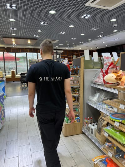 A man walks away from the camera down an aisle in a brightly lit convenience store. He is wearing a black t-shirt with white Cyrillic lettering that reads "Я НЕ ЗНАЮ" (Ya ne znayu), which translates to "I don't know." He is also wearing black pants. The store aisle is lined with shelves displaying various products, including drinks, toys, and household items. The flooring is made of light-colored tiles. In the background, through a window, cafe seating can be seen. The scene appears to be indoors, possibly a gas station convenience store, given the typical offerings visible on the shelves and the overall ambiance.