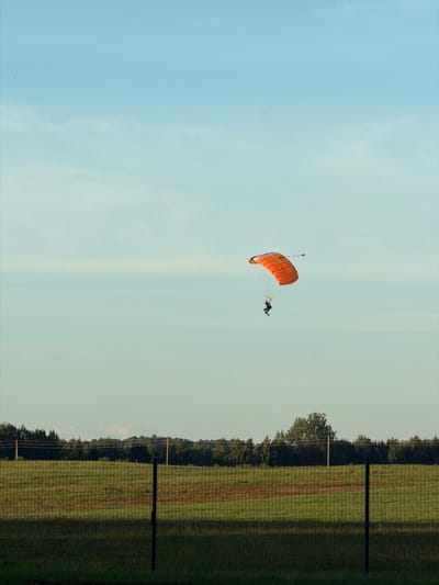 The image depicts a paraglider in mid-air, descending against a pale blue sky with faint wispy clouds. The paraglider, primarily orange with some black accents, is visible in the upper right quadrant of the frame. A person in dark clothing is suspended beneath the parachute. Below the sky, a line of dark green trees forms a distant horizon. In the foreground, a green, checkered field is visible, separated from the viewer by a dark wire fence with vertical posts. The overall scene suggests an open, rural area, possibly near an airfield or a designated paragliding site. The lighting indicates it is likely daytime, with soft sunlight casting shadows on the field. There is no specific indication of the city of Limbaži, Latvia, within the visual elements of the image.