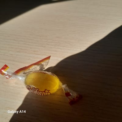 The image shows a yellow lollipop, wrapped in clear plastic with red and white stripes at the twisted ends. The lollipop is on a light-colored wooden surface with a dark shadow partially obscuring the scene. "California" is printed on the lollipop wrapper. The location is Unknown in the city of Al-Mawasi. The device used to capture the image is a Galaxy A14.