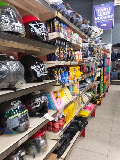 This image presents an interior view of an aisle within a retail store, likely specializing in motorcycle accessories and automotive products, set in Sindanglaya, Indonesia. The scene is well-lit with artificial lighting.
Multiple shelves, constructed from wood and metal, extend through the foreground and midground, showcasing a diverse range of merchandise. Dominating the lower and upper shelves are numerous motorcycle helmets of various designs and colors, many still protected by clear plastic coverings. Visible helmet brands include "KYT," "BMC," and "HJH."
The shelves are densely packed with other products, including aerosol cans of sprays (possibly for vehicle maintenance or cleaning), various packaged items such as gloves, cleaning cloths, and small automotive accessories. These items are displayed in bright packaging, featuring colors like yellow, blue, pink, and red. Handwritten price tags and product labels are visible on some shelves, indicating specific items like "KIT."
In the background, further shelving units display what appear to be bags or backpacks. A prominent blue banner hangs from the ceiling in the upper right. The banner features Indonesian text: "PASTI LEBIH MURAH!!" (Definitely Cheaper!!) and "MAHAL, UANG KEMBALI" (Expensive, money back), serving as a promotional advertisement for the store.
The image provides a snapshot of a retail environment, organized for customers to browse a wide selection of products, without any individuals present.