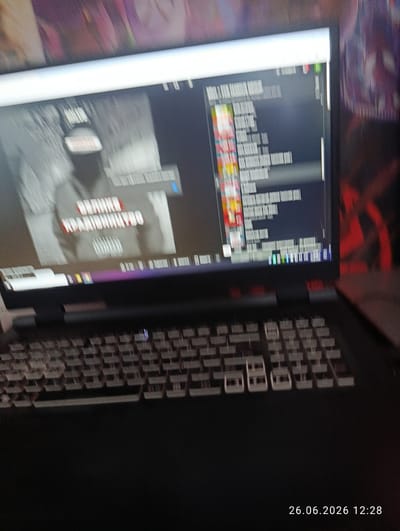 The image shows a laptop computer placed on a keyboard. The laptop's screen displays a video with Russian text. The video appears to be a music video or a documentary, with a person wearing a cap and sunglasses in the foreground. The text on the screen is blurry, but some phrases are partially visible, such as "ВЕЛИКОЕ КРАЙНЕЕ" and "PAID". The scene is lit by the screen's glow, and the background is dark with some blurry colorful shapes. A timestamp in the bottom right corner indicates "26.06.2026 12:28". The location is identified as Novovolynsk, Ukraine. The overall impression is a candid shot of someone using a laptop, possibly for entertainment or work.